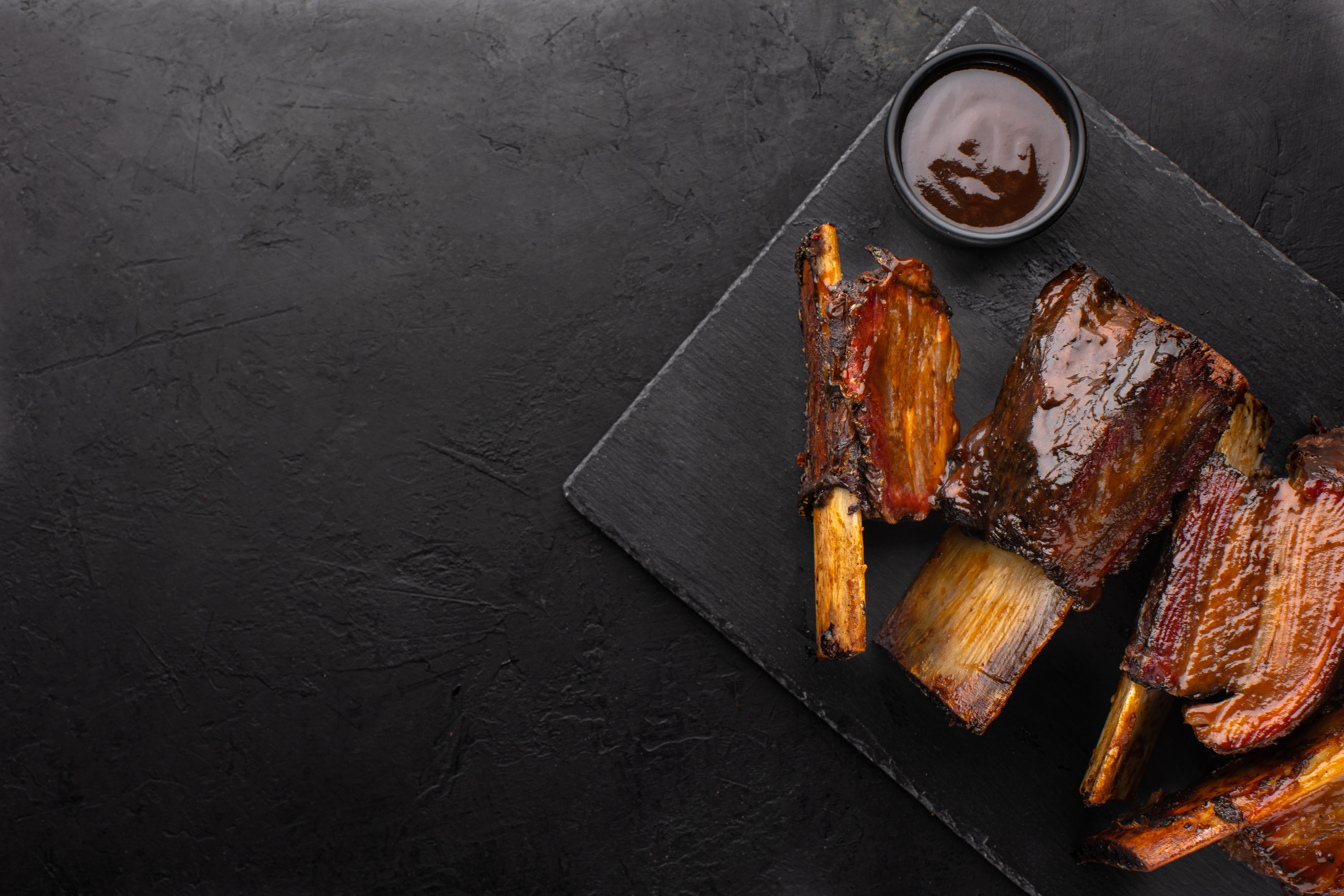 Smoked beef ribs with bone and barbecue sauce on black slate board, top view, copy space. Roasted beef ribs.